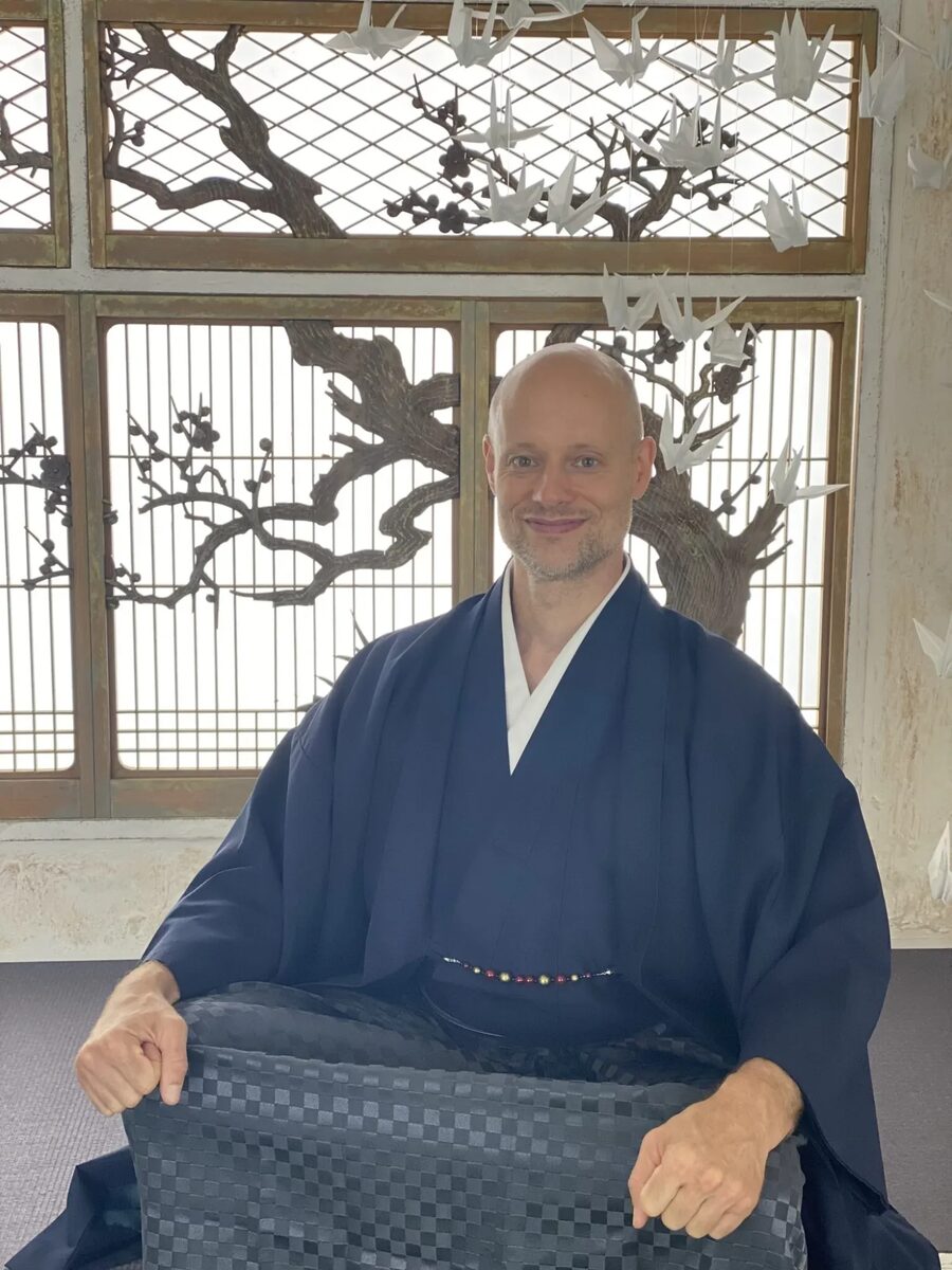 Rob Daoust in a dark blue kimono, seated in front of a lattice window with a plum tree design